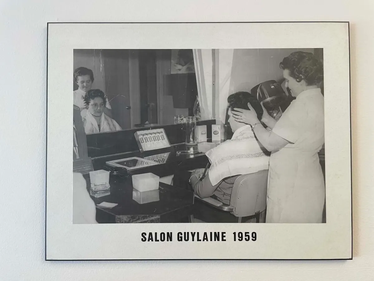 Photo d’archive en noir et blanc du Salon Guylaine en 1959 — deux coiffeuses au travail devant les miroirs du salon.